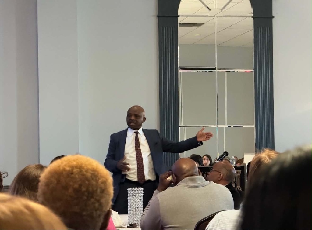 Photo of an Dr. Mubenga standing at the front of a room speaking to an audience seated at tables. He gestures with one hand while addressing attendees, with microphones and event equipment visible nearby. The setting appears to be an indoor meeting or presentation space with tall mirrors and columns in the background.