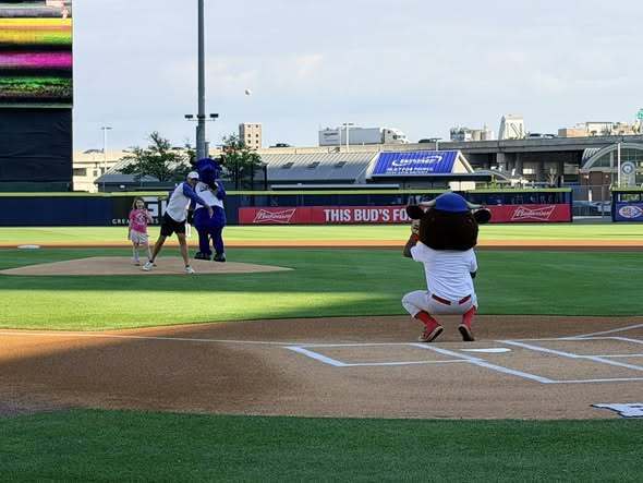 Child throws a ceremonial first pitch on a baseball field while a catcher kneels at home plate and a team mascot stands nearby. Stadium seating and scoreboard are visible in the background.
