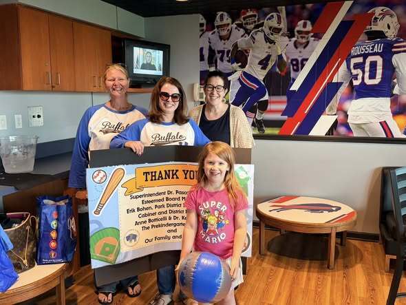 Group standing indoors holding a ‘Thank You’ sign with a child holding a blue ball in the foreground; Buffalo football wall graphics and seating area visible in the background.