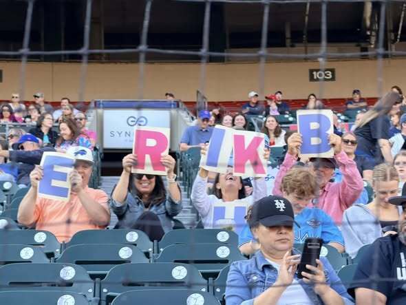 Group of people seated in a stadium holding letter signs that spell ‘ERIKA B’ while others watch and take photos