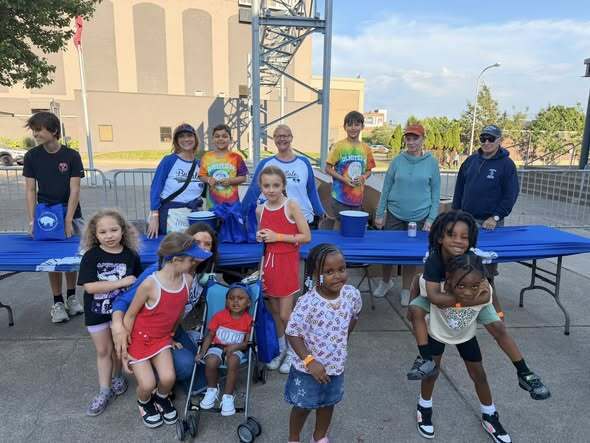 Outdoor community event with children and adults gathered around tables with blue bags and supplies in an open paved area.
