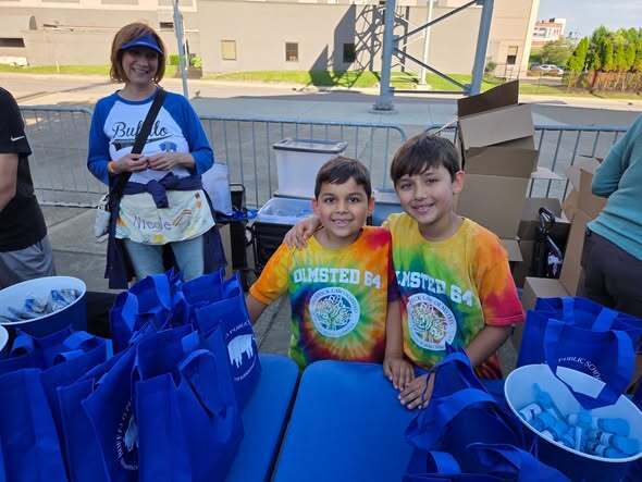 Two children standing behind a table filled with blue bags and supplies at an outdoor event, with additional materials and boxes nearby.