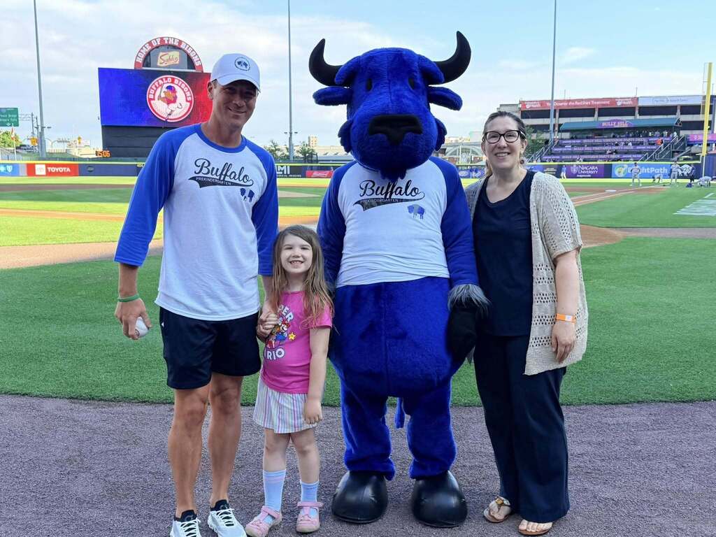 Group of people posing on a baseball field with a large blue bison mascot, with the Buffalo Bisons scoreboard and stadium seating in the background.