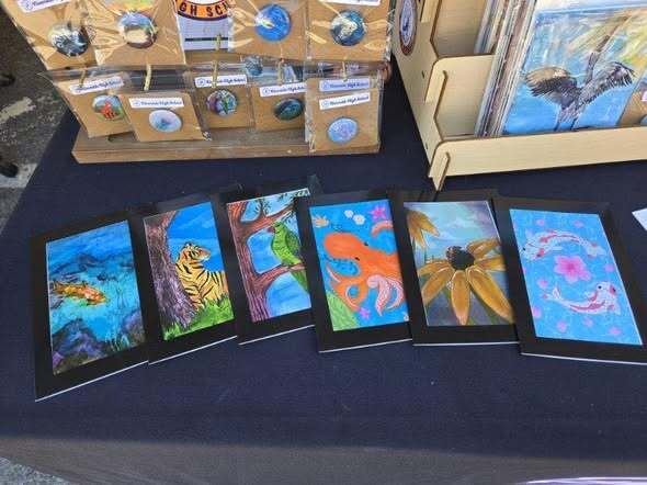 “Table display of student-made art prints and small items featuring colorful designs of animals and plants, with additional packaged items arranged on stands behind.”