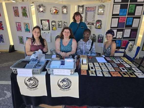 Group seated behind an outdoor display table with art-making materials and handmade items, with student artwork panels exhibited in the background.