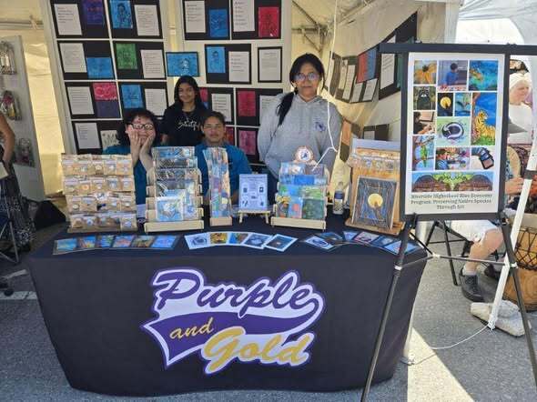 “Group standing behind an outdoor booth displaying student artwork, prints, and packaged items, with a sign reading ‘Purple and Gold’ on the table and an art display board to the side.”