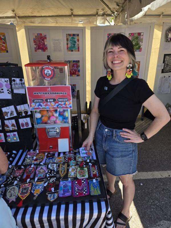 “Outdoor arts display table featuring handmade jewelry, a red ‘mystery earrings’ vending machine, and student artwork panels in the background.”