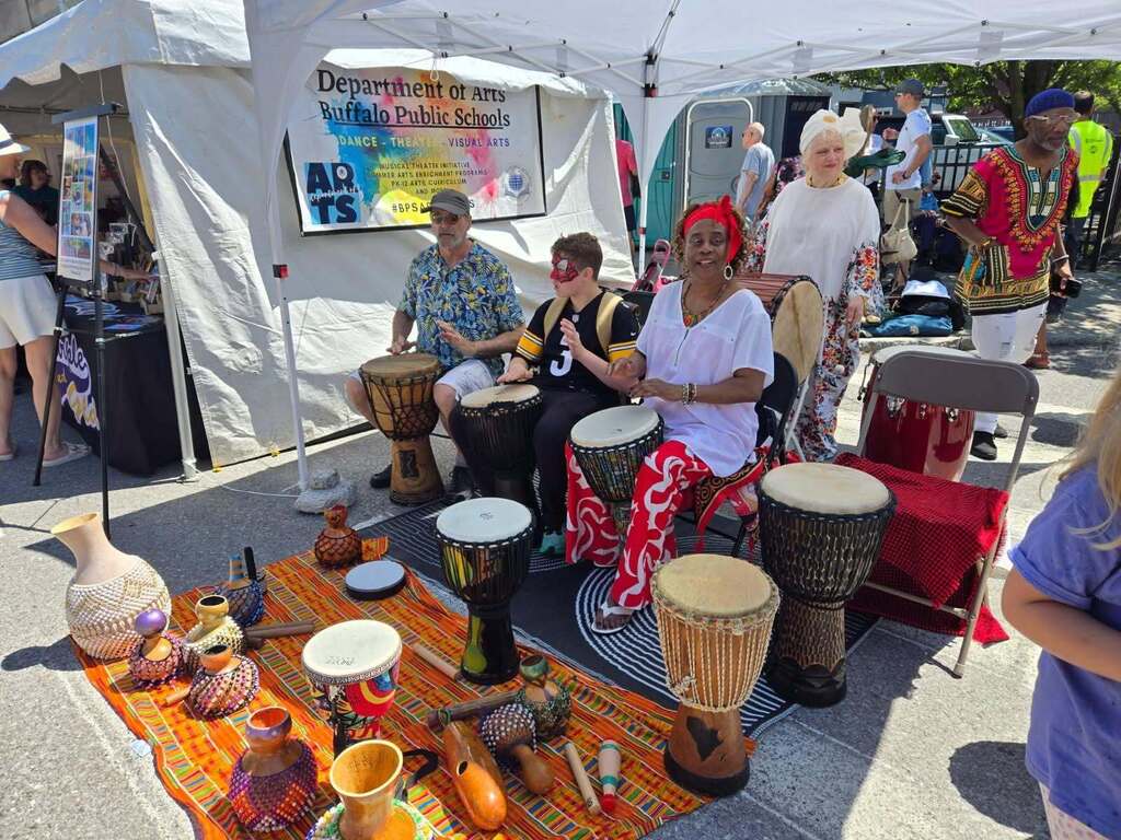 Buffalo Public Schools Department of Arts tent at an outdoor event, with participants playing hand drums and percussion instruments displayed on a table in front.