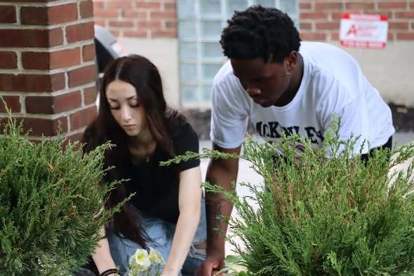 “Two students planting flowers in a landscaped garden bed near a brick building, with small shrubs and soil visible in the foreground.”