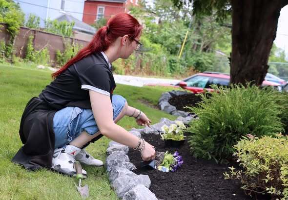 “Student kneeling in a garden bed bordered with stones, placing flowers into soil alongside shrubs and grass in a landscaped outdoor area.”