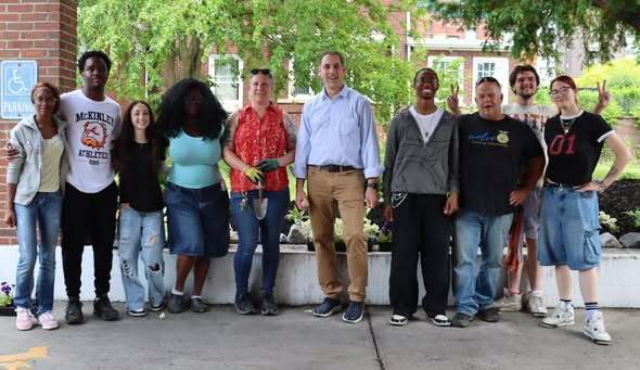 “Group of students and adults standing together outside near a building and garden area, with plants and trees in the background.”