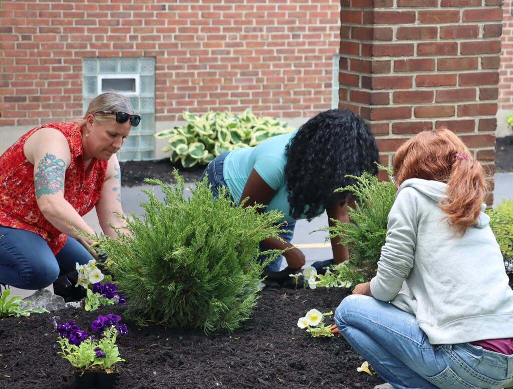 “Students and an adult plant flowers in a garden bed near a building.”