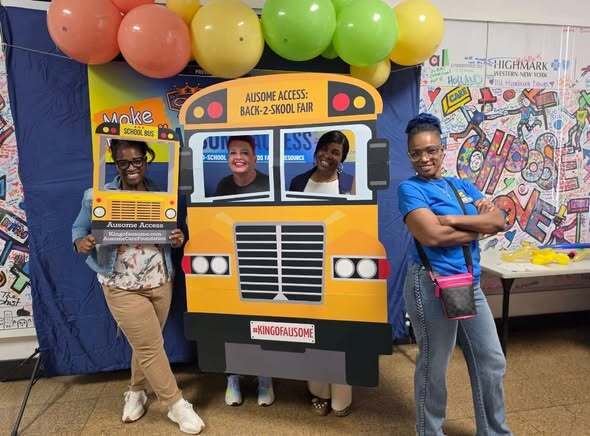 “Group posing with school bus-themed photo props at the Ausome Access Back-2-Skool Fair. The display features colorful balloons, event branding, and a community mural wall in the background.”