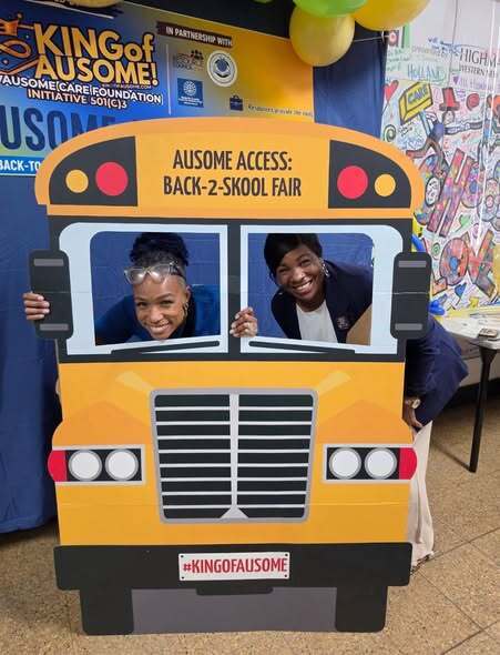 “Attendees pose behind a school bus-themed photo prop at the Ausome Access Back-2-Skool Fair. The display is decorated with colorful balloons and event signage from the King of Ausome initiative.”