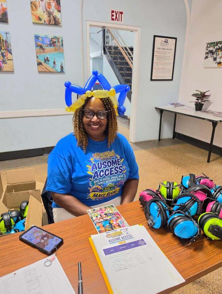 Resource fair registration table with stacks of colorful noise-reducing headphones, sign-in sheets, informational materials, and a balloon hat. Community event posters and displays are visible in the background.”