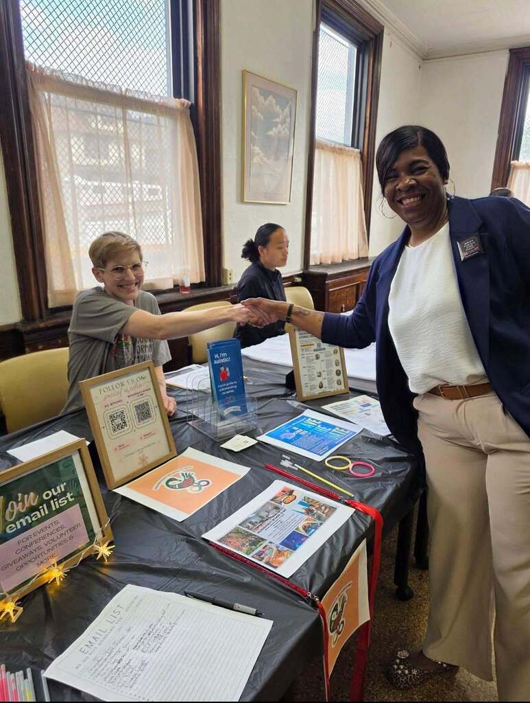 Community resource fair information table with brochures, sign-up materials, and informational displays. Two people shake hands across the table while another attendee sits behind the display area in a meeting room with large windows.”