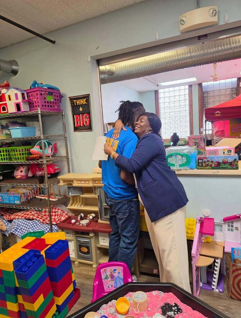 “Two attendees greet each other inside a classroom-style activity space furnished with toy shelves, block-building materials, dollhouses, and sensory play items. Resource fair supplies and activity stations are visible throughout the room.”
