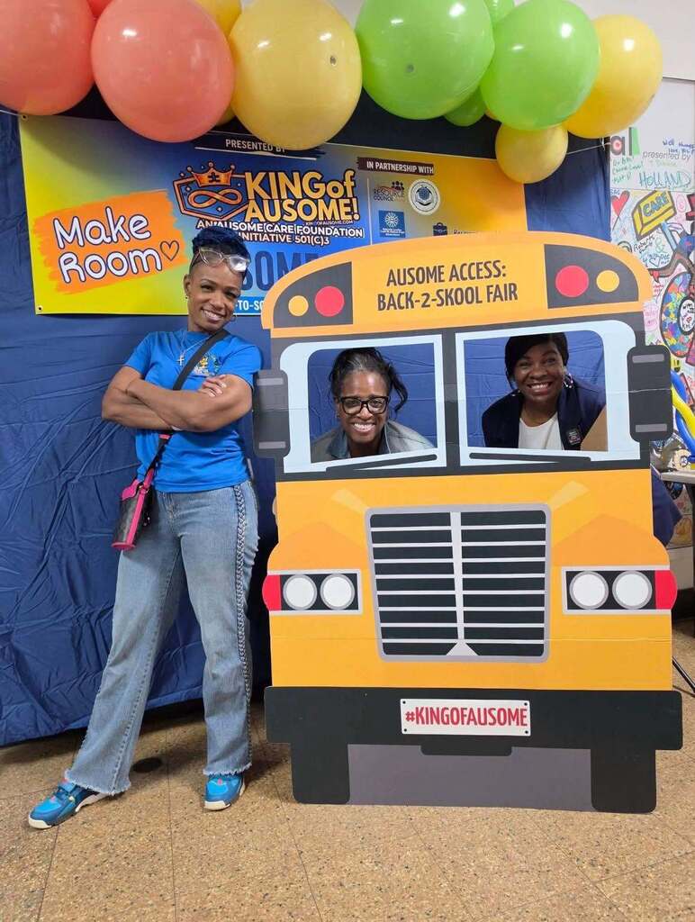 Back-to-school resource fair display featuring a large school bus photo prop, colorful balloons, and event signage reading ‘Ausome Access: Back-2-Skool Fair.’ Three attendees pose with the bus display in front of community resource organization banners