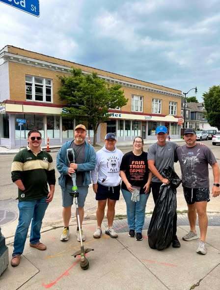 “Six community volunteers pose on a city sidewalk with litter-collection equipment and a filled trash bag during a neighborhood beautification project. Commercial buildings and street signs are visible in the background.”