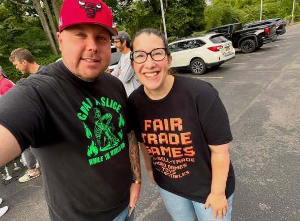 “Two volunteers pose in a parking lot during a community cleanup event. Additional participants and parked vehicles are visible in the background.”