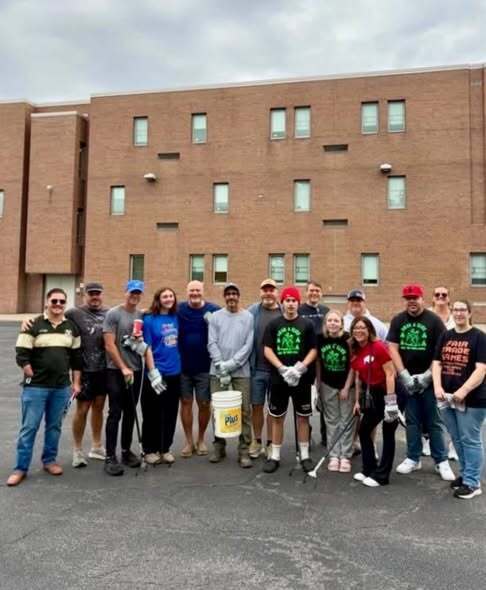 “Group of community volunteers standing together outside a brick building during a neighborhood beautification project. Several participants hold litter-collection tools, gloves, and buckets used for cleanup activities.”