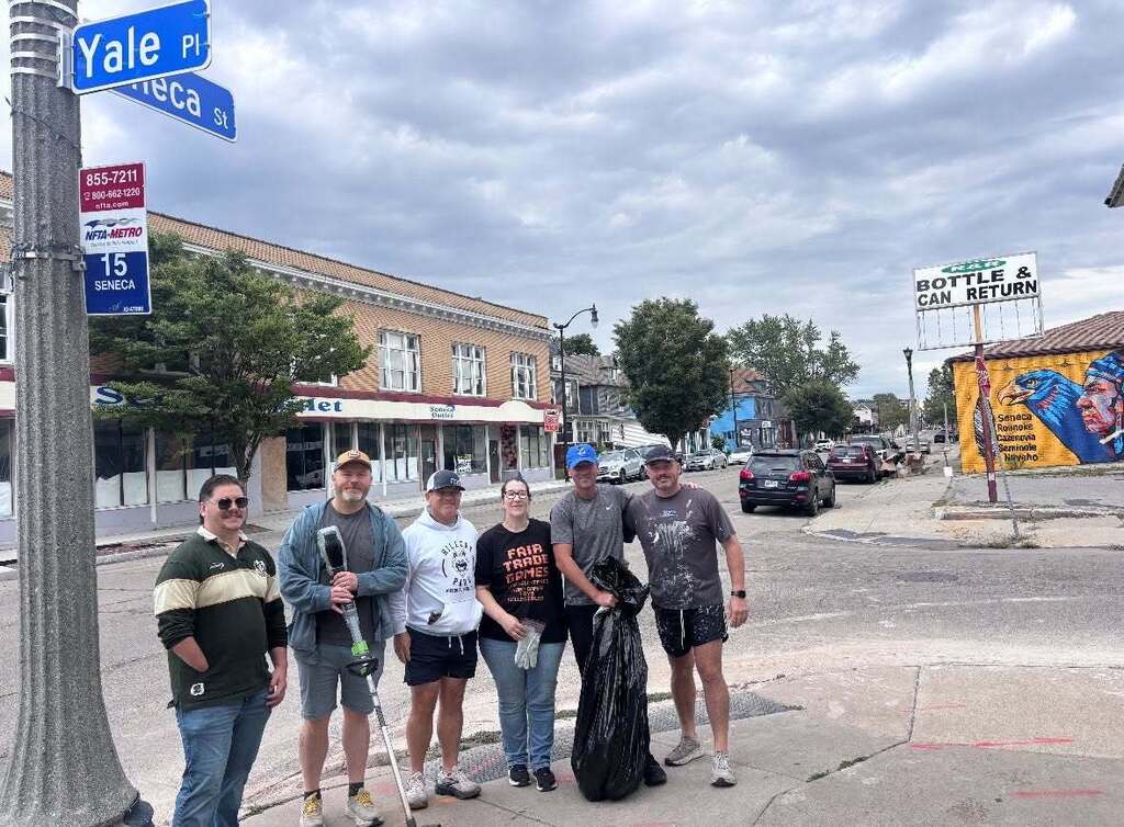 “Group of community volunteers standing at the corner of Yale Place and Seneca Street during a neighborhood cleanup. The group gathers with trash bags and litter-collection tools near local businesses and a colorful mural.”