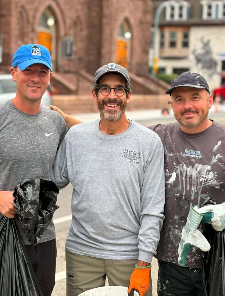 “Three community volunteers pose outdoors holding cleanup supplies, including trash bags and work gloves, in front of a brick building. One volunteer wears protective gloves while participating in a neighborhood cleanup effort.”