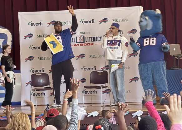 “Students raise their hands while participating in a financial literacy assembly. Presenters stand on stage in front of a backdrop displaying Buffalo Bills, M&T Bank, EverFi, and Financial Literacy Playbook logos, alongside a blue buffalo mascot.”