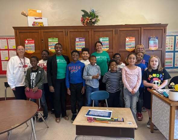 “Students and volunteers pose together in a classroom during a financial literacy and education outreach visit. Classroom posters, student desks, and learning materials are visible in the background.”