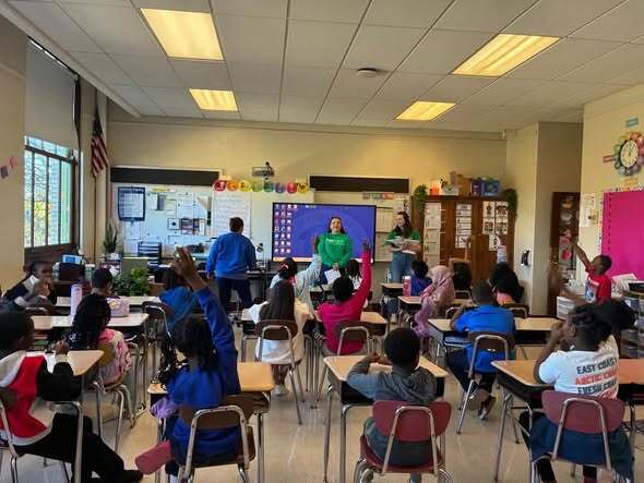 “Volunteer leads a classroom activity with elementary students while standing near an interactive display screen. The classroom includes educational posters, student desks, and learning materials.”