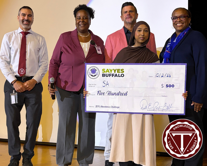 “Student stands with school and district representatives while holding a ceremonial Say Yes Buffalo attendance award check for $500 during a school recognition event. A school logo appears in the lower-right corner.”
