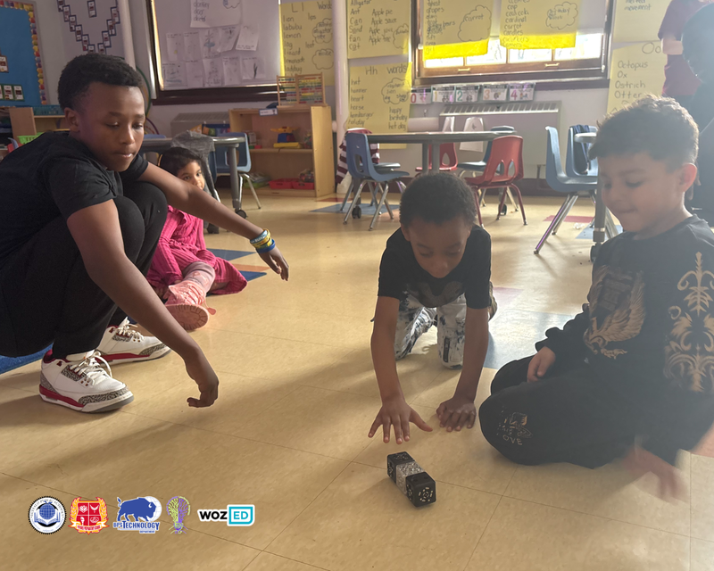 An STL guiding two young students using robotics on the floor of a classroom