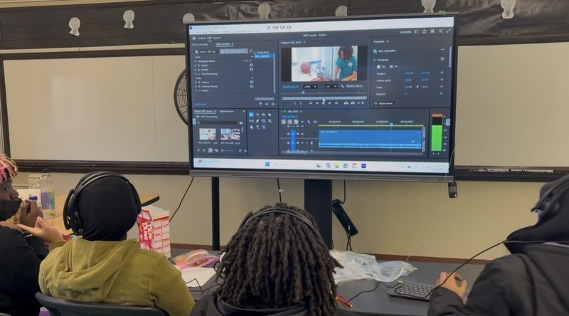 Students sit at a classroom table wearing headsets, facing a large display that shows video editing software with a timeline and preview window. Computers, a keyboard, and editing materials are visible as the group works together on a film project.