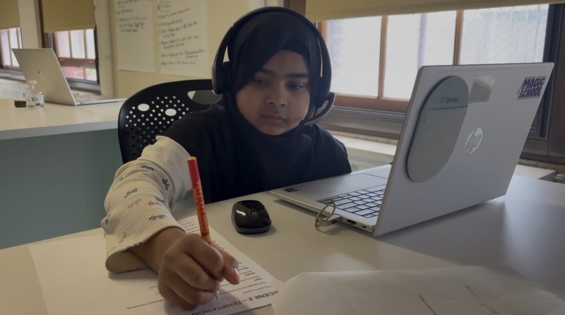 A student sits at a classroom desk wearing a headset, working on a laptop while writing on a printed worksheet with a pencil. A computer mouse and papers are on the desk, with windows and classroom materials visible in the background.