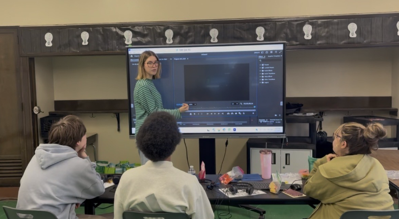 A student sits at a classroom desk wearing a headset, working on a laptop while writing on a printed worksheet with a pencil. A computer mouse and papers are on the desk, with windows and classroom materials visible in the background.