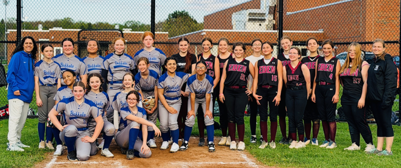 Group photo of two softball teams and coaches standing together on a softball field. Players are wearing uniforms from opposing teams and are lined up along the baseline in front of a chain‑link backstop. A school building and athletic field fencing are visible in the background under a cloudy sky.