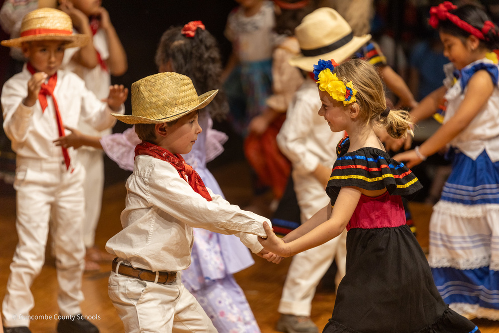 A boy in a straw hat extends his hands to a girl with a flower headband as they two dance on stage.