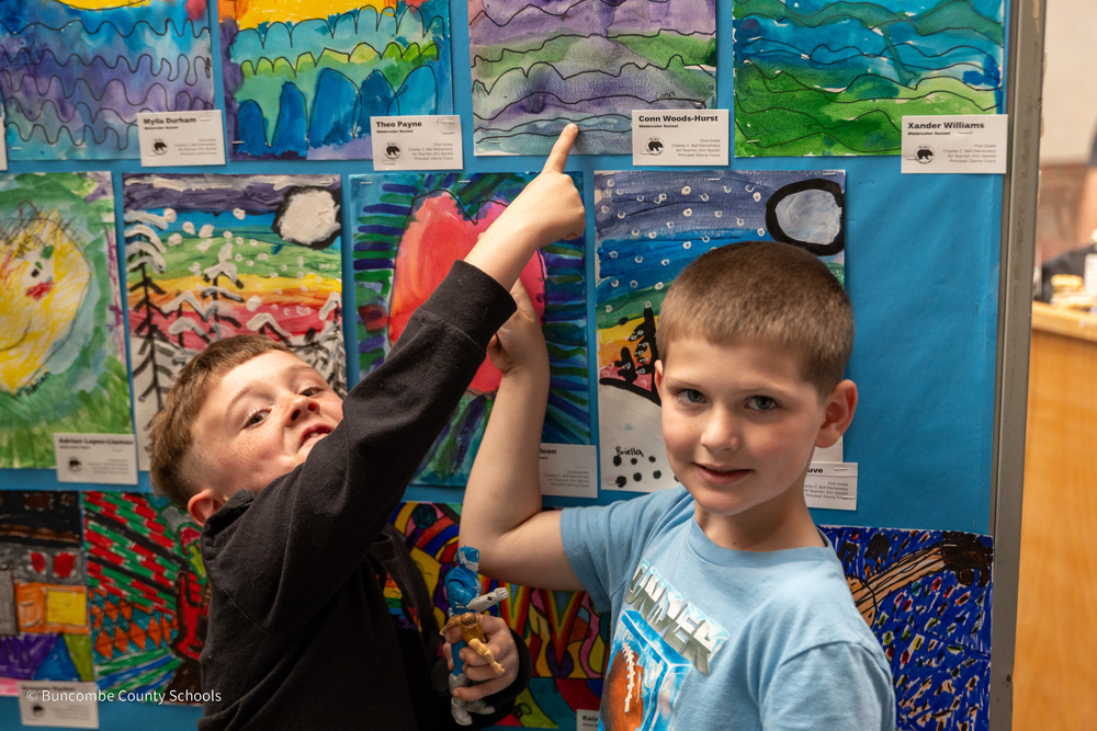 Two students stand in front of a wall of artwork in the mall. They are pointing to the pieces they created.