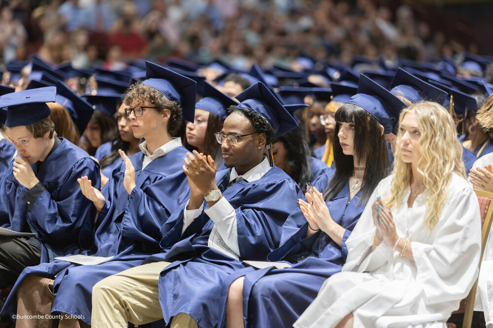 Graduates dressed in blue caps and gowns seated in an arena, applauding.