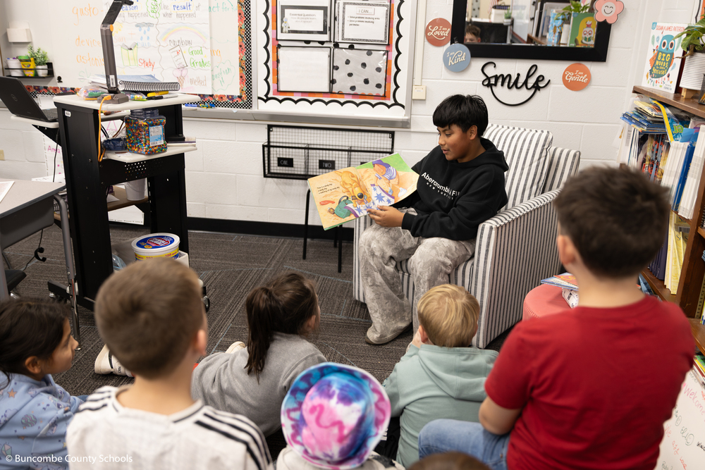 Middle school student reading a book to a small group of third graders.