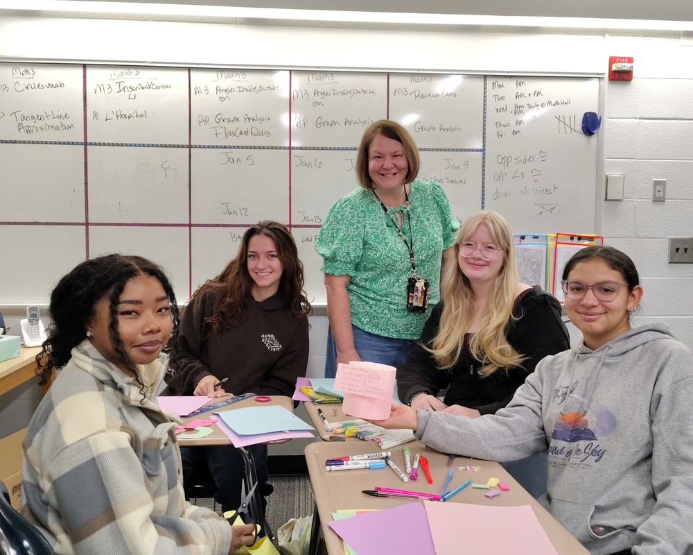 Mrs. Franklin stands in front of her white board with two students on either side, seated at desks.
