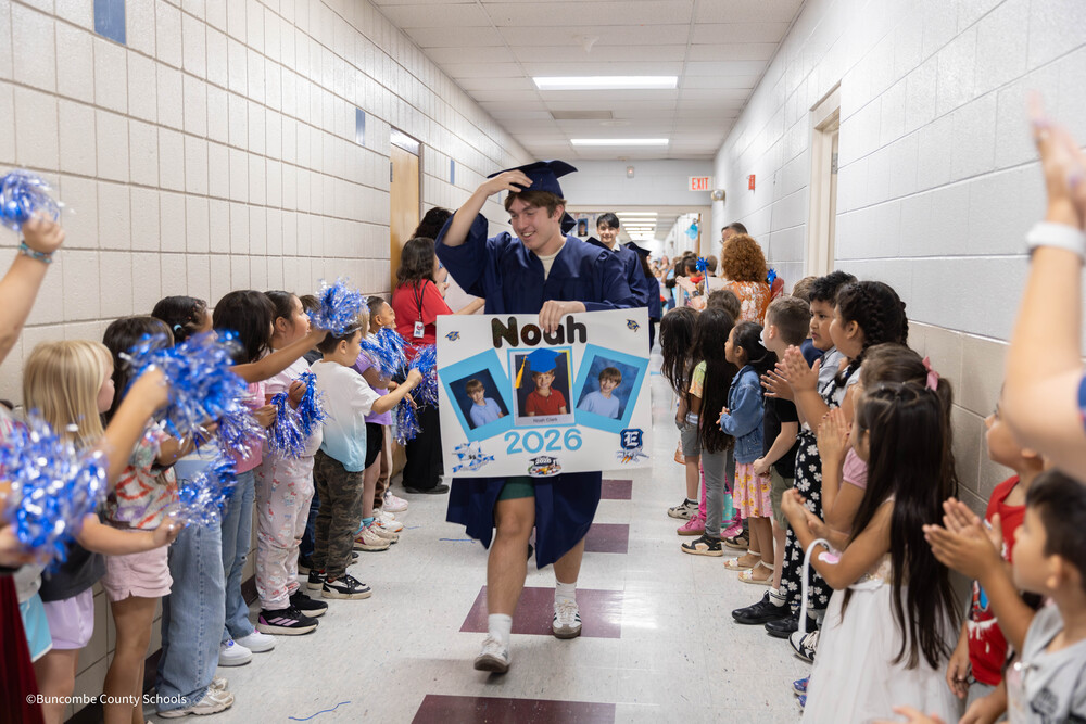 A senior in blue cap and gown walks down a hallway lined with elementary students.