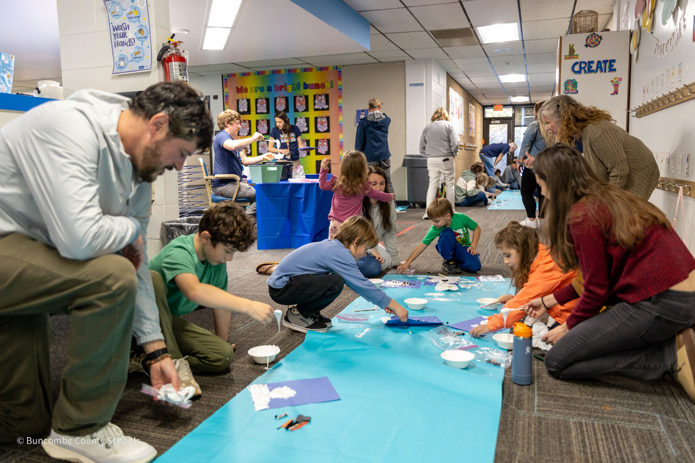 Parents and students sit on the floor around a long piece of blue construction paper. They are decorating it with  a shaving cream solution that resembles snow.