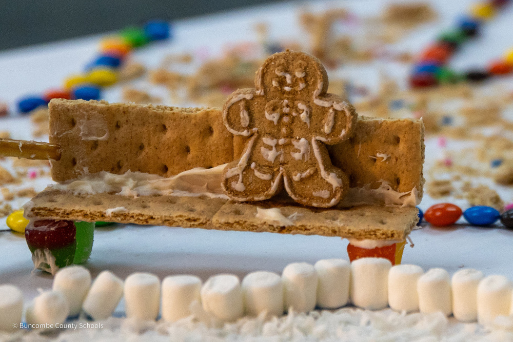 A gingerbread man cookie sits on a bench made of graham crackers. A ring of mini-marshmallows is below the bench.