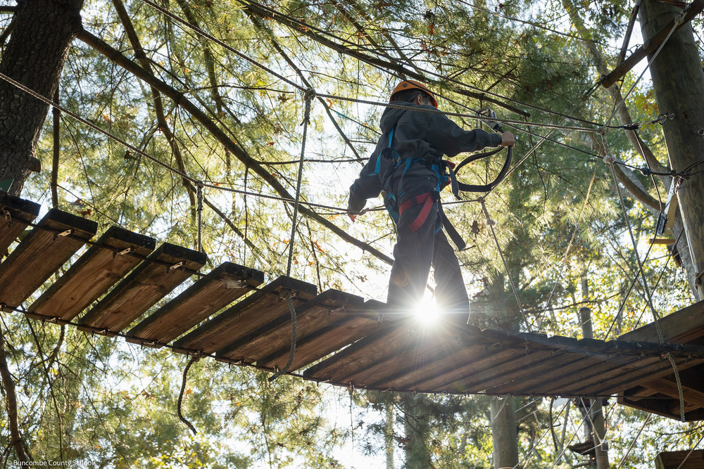 A student walks across a rope bridge with the sun shining through.