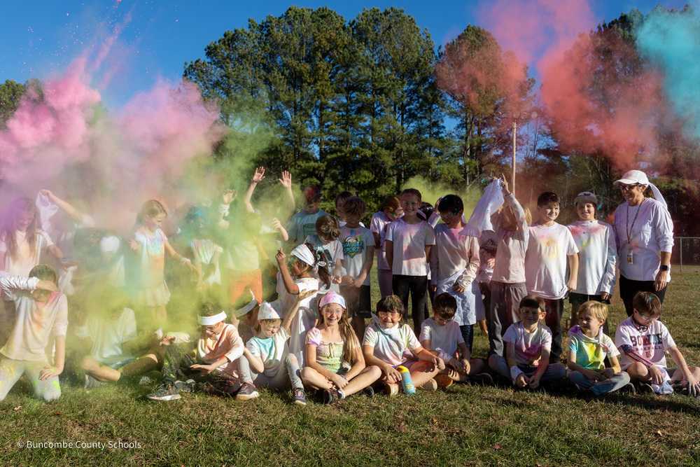 A group shot of students who participated in the color run. Puffs of pink, yellow, blue and red colored powder is floating in the air.