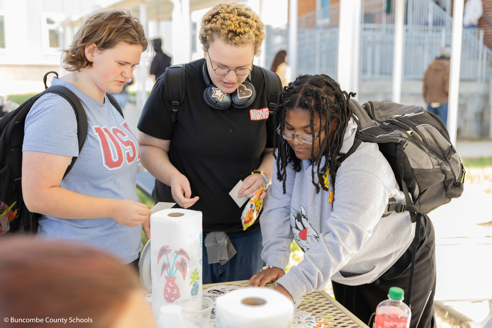 Three students looking down at something lying on the picnic table. 