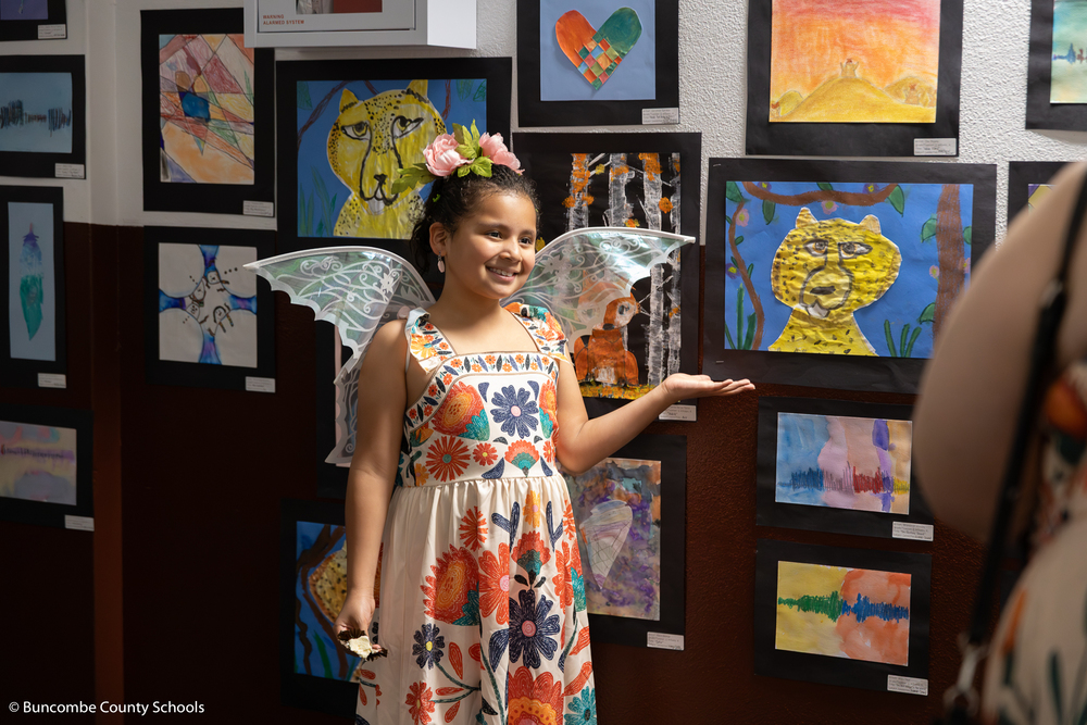 Girl wearing a dress and fairy wings, pointing at her artwork on the wall as her mother takes a photo. 