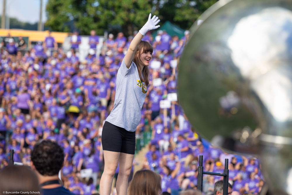 Drum major Ever van Rijssen stands elevated on the football field directring the band. A crowd of people are behind her in the stands. A tuba is visible in front of her.