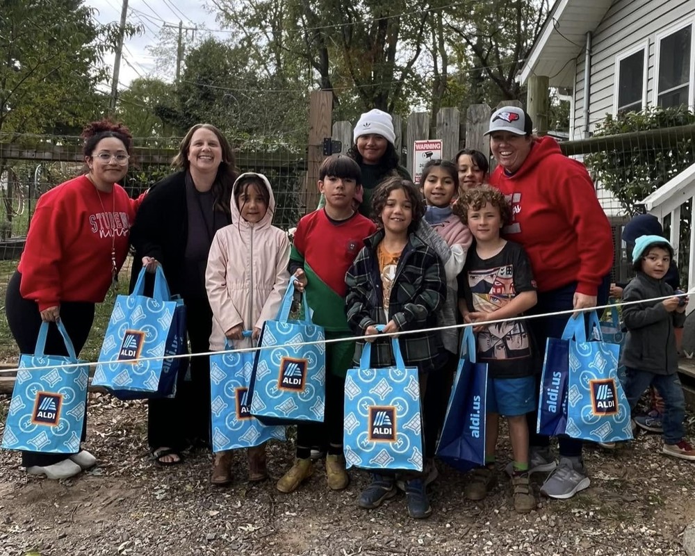 Emma Elementary staff members stand in a line with students outside a family's home. Everyone is holding a blue plastic Aldi bag filled with food and supplies.